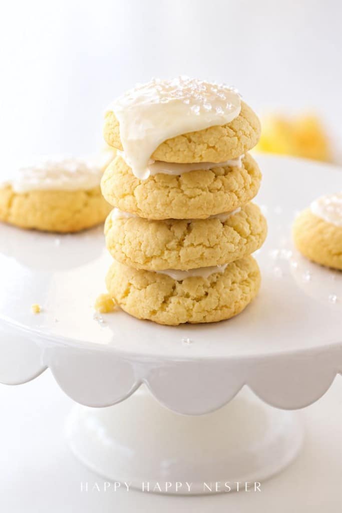 A stack of four soft, yellow cookies made from a delicious lemon sugar cookie recipe with white icing and sprinkles sits on a white scalloped cake stand. More cookies are visible in the background, all in a bright, light-filled setting.