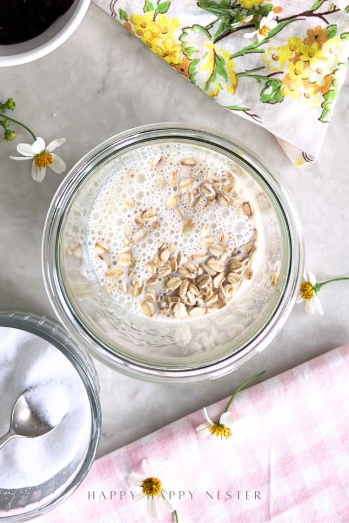 A glass bowl of oats soaking in milk sits on a marble surface, ready for an overnight oats recipe with yogurt. Nearby are a bowl of sugar, small white flowers, a yellow floral napkin, and a pink checked cloth.