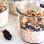 A glass jar filled with an overnight oats recipe with yogurt, blackberries, and granola sits on a white surface, with two more jars and loose blackberries in the background.