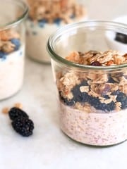 A glass jar filled with an overnight oats recipe with yogurt, blackberries, and granola sits on a white surface, with two more jars and loose blackberries in the background.