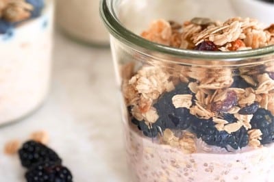 A glass jar filled with an overnight oats recipe with yogurt, blackberries, and granola sits on a white surface, with two more jars and loose blackberries in the background.