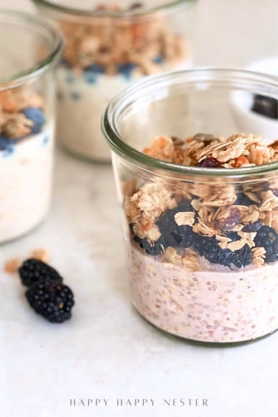 A glass jar filled with an overnight oats recipe with yogurt, blackberries, and granola sits on a white surface, with two more jars and loose blackberries in the background.