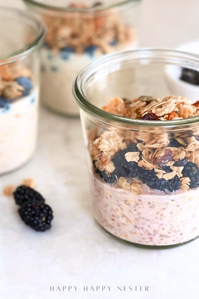A glass jar filled with an overnight oats recipe with yogurt, blackberries, and granola sits on a white surface, with two more jars and loose blackberries in the background.