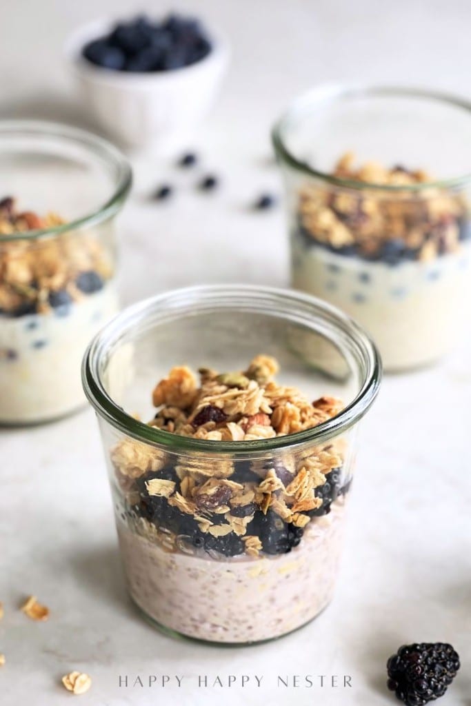 Three glass jars filled with an overnight oats recipe with yogurt, topped with granola and blueberries, are arranged on a light surface. A small white bowl with extra blueberries sits in the background.