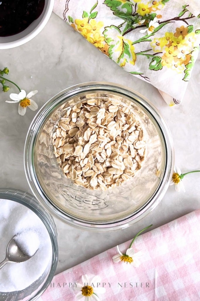 A glass bowl filled with rolled oats sits on a marble surface, perfect for an overnight oats recipe with yogurt, surrounded by a bowl of sugar with a spoon, a floral napkin, pink checkered cloth, small white flowers, and part of a dark jam bowl.
