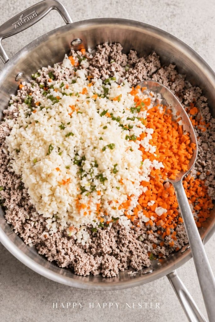 A skillet filled with cooked ground meat, topped with piles of finely chopped cauliflower and carrots, with a large metal spoon resting on the mixture.