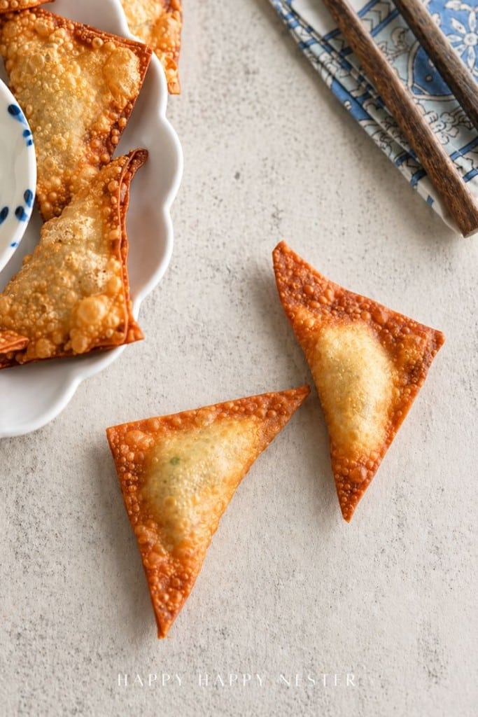 Two golden-brown, crispy wontons rest on a light surface near a plate of more wontons, showcasing the perfection of this fried wonton recipe, with chopsticks and a blue patterned napkin nearby.