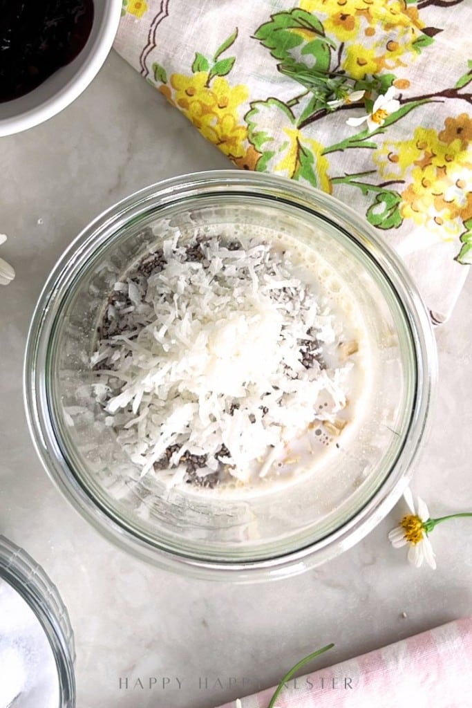 A glass bowl filled with shredded coconut, chia seeds, and milk sits on a light surface next to a yellow floral napkin, small daisies, and other ingredients.