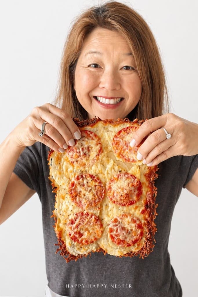 A smiling woman holds up a rectangular, cheesy baked dish topped with sliced tomatoes, showcasing her onion ring pizza recipe. She wears a dark shirt and stands against a plain white background.