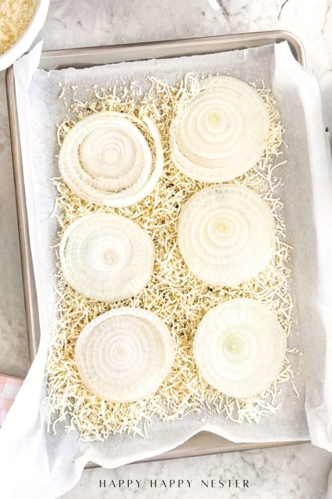 Six large, raw onion slices placed on shredded cheese on a parchment-lined baking sheet, ready for baking. The scene is set on a light marble surface.