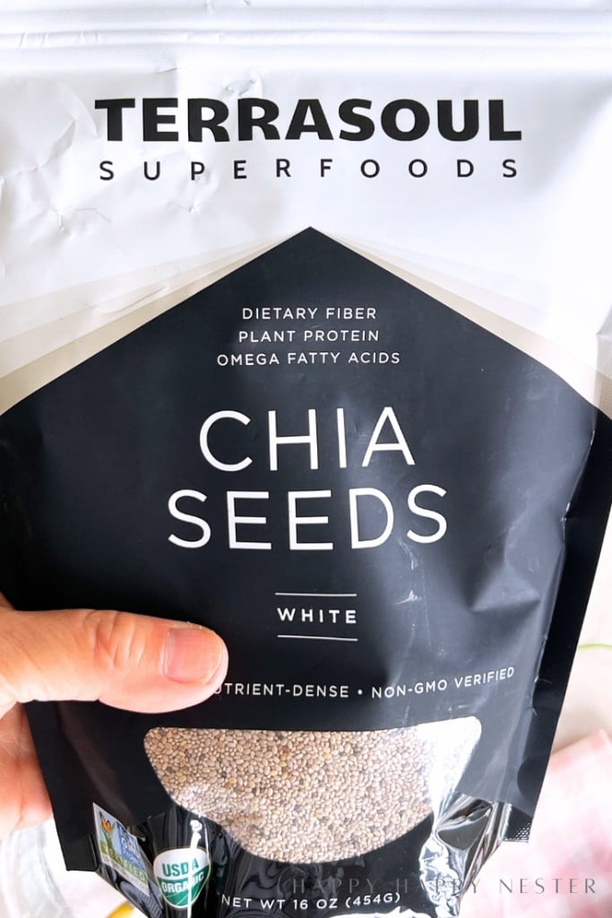A hand holds a bag of Terrasoul Superfoods White Chia Seeds. The package highlights dietary fiber, plant protein, omega fatty acids, and is labeled nutrient-dense, non-GMO verified, and USDA organic.