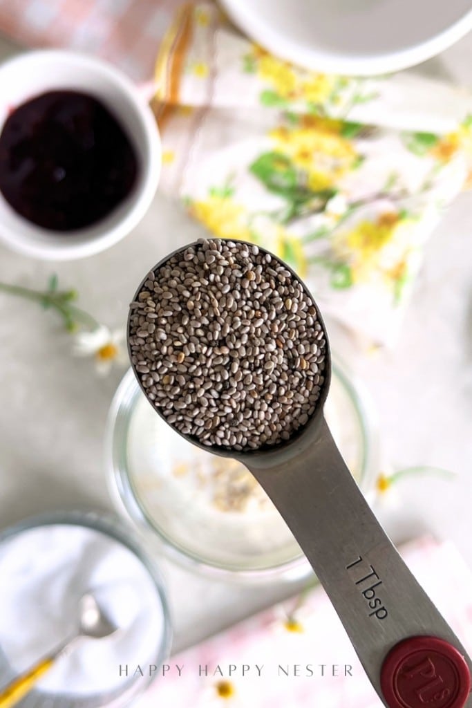 A close-up of a tablespoon filled with chia seeds above a countertop with various bowls and a floral-patterned cloth in the background.