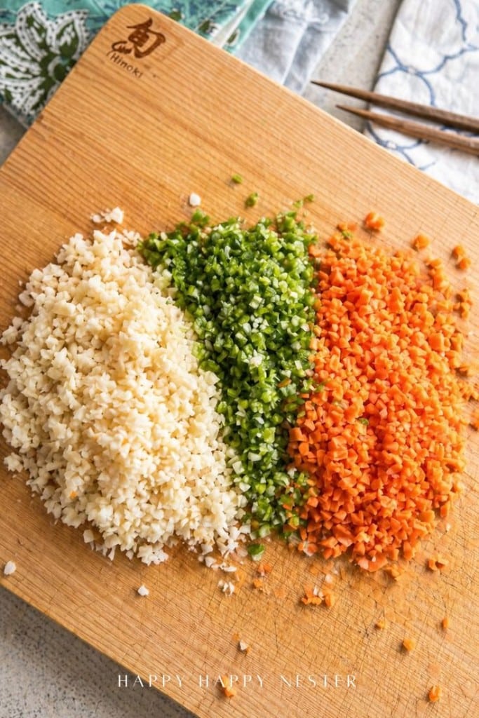 Finely chopped cauliflower, green bell pepper, and carrot are arranged in neat piles on a wooden cutting board with a knife and chopsticks visible nearby.