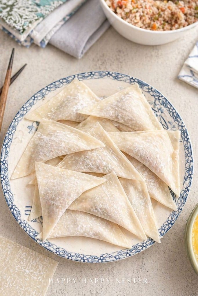 A decorative plate holds several uncooked, triangular dumplings wrapped in thin dough, dusted lightly with flour, ready for your favorite fried wonton recipe. Surrounding the plate are small bowls of ingredients and stacked napkins.