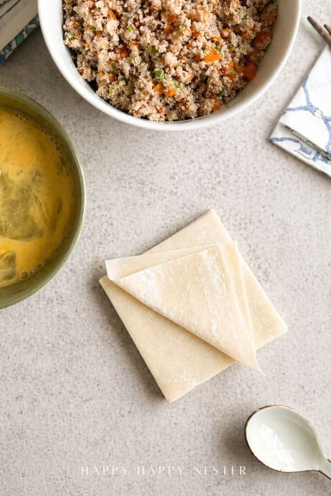 Top-down view of a bowl of mixed couscous, a bowl of beaten eggs, a small stack of square wonton wrappers, a white spoon, and a napkin on a light-colored surface.