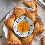 A plate of golden, crispy wontons—made with a classic fried wonton recipe—arranged around a small bowl of sweet chili dipping sauce, with a bowl of grain salad and napkins in the background.