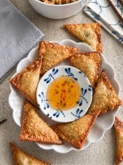 A plate of golden, crispy wontons—made with a classic fried wonton recipe—arranged around a small bowl of sweet chili dipping sauce, with a bowl of grain salad and napkins in the background.