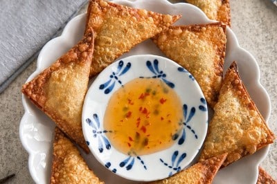 A plate of golden, crispy wontons—made with a classic fried wonton recipe—arranged around a small bowl of sweet chili dipping sauce, with a bowl of grain salad and napkins in the background.