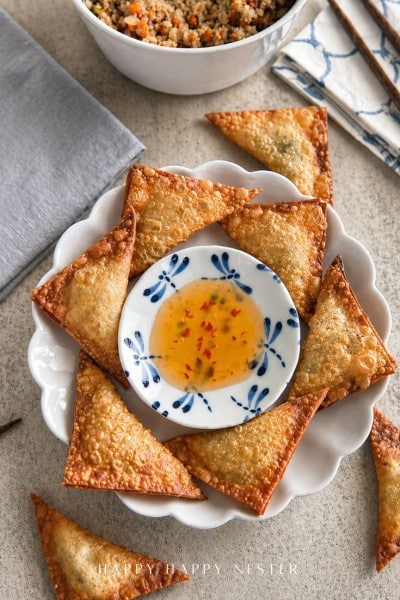 A plate of golden, crispy wontons—made with a classic fried wonton recipe—arranged around a small bowl of sweet chili dipping sauce, with a bowl of grain salad and napkins in the background.