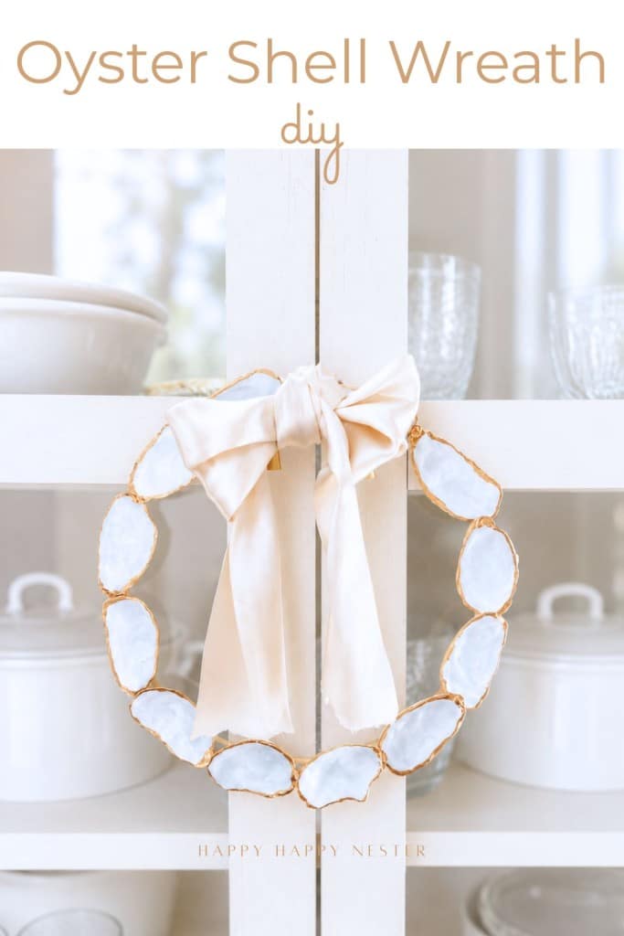 A decorative oyster shell wreath diy, featuring white shells with gold edges and a large cream-colored bow, hangs on a cabinet filled with glassware and kitchenware. Text reads "Oyster Shell Wreath diy.