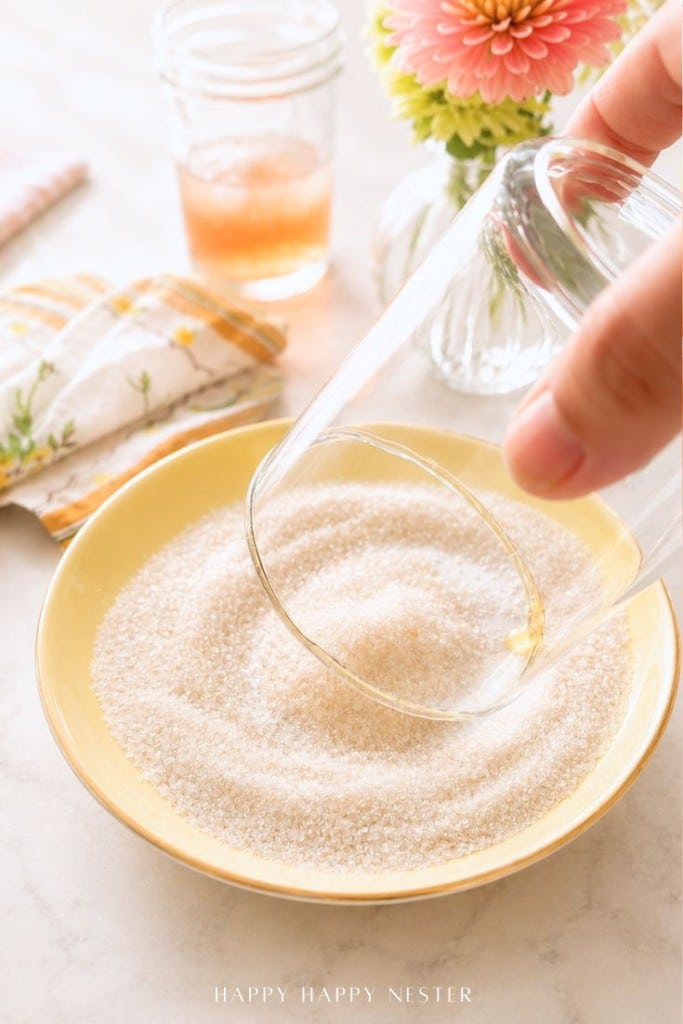 A hand presses a glass into a bowl of sugar on a light countertop, with a floral napkin, a jar of pink liquid, and a vase with a pink flower in the background.