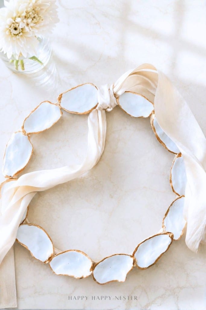 A decorative oyster shell wreath DIY, crafted with white shells edged in gold and tied with a cream ribbon, is displayed on a white marble surface beside a glass vase filled with white flowers.