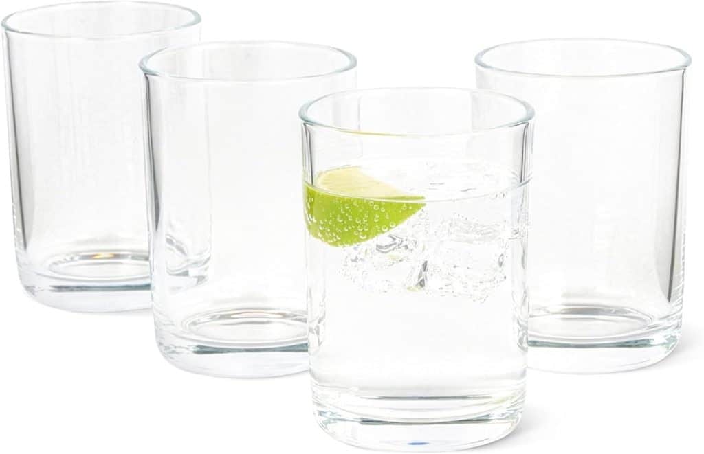 Four clear glass tumblers are arranged in a group. Three are empty, and one in front contains ice, sparkling water, and a lime wedge—perfect for serving your favorite ginger beer mocktail recipe.