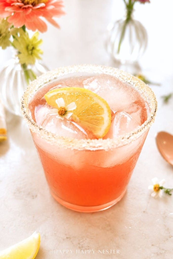 A glass of pink iced cocktail with a sugared rim, garnished with a lemon slice and small white flower, sits on a light surface with flowers and a spoon nearby.