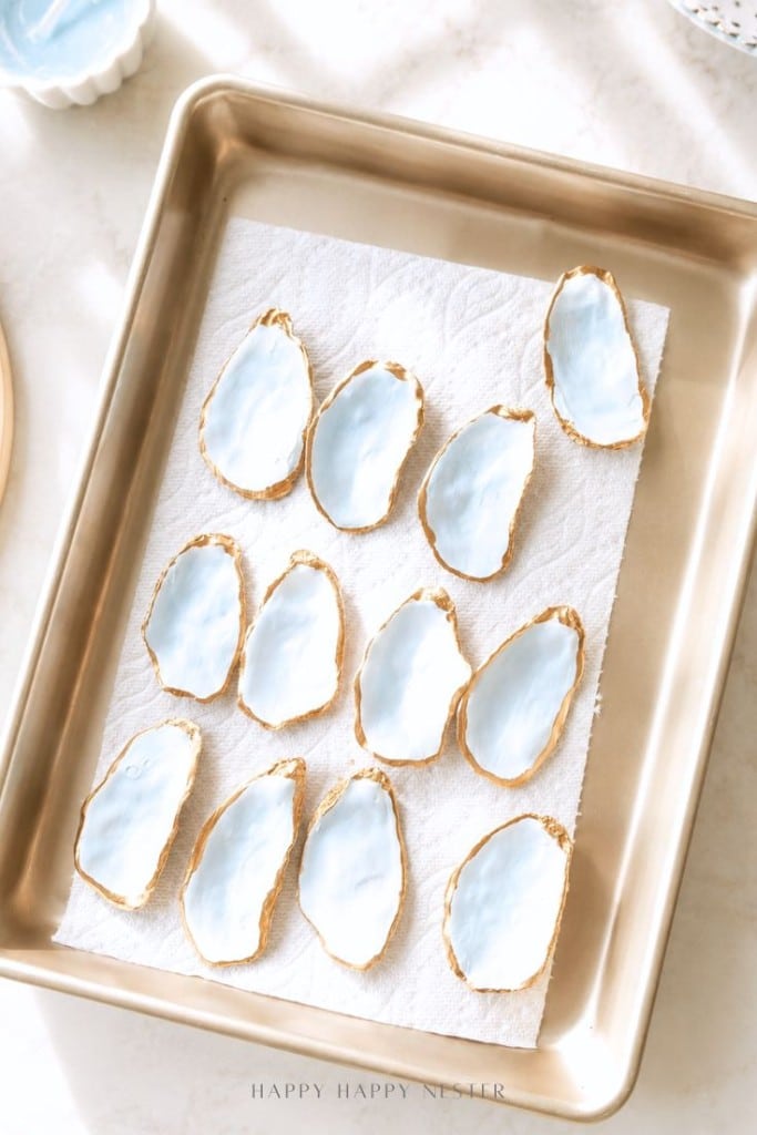 A baking tray lined with paper towels holds twelve oyster shells, each painted light blue inside with gold edges, arranged neatly in rows on a light-colored surface.