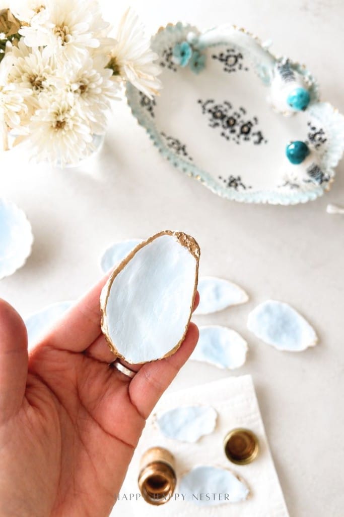 A hand holds a decorative oyster shell painted light blue with a gold edge. Several similar shells, a small brass container, a white flower bouquet, and a decorative tray are visible in the background.