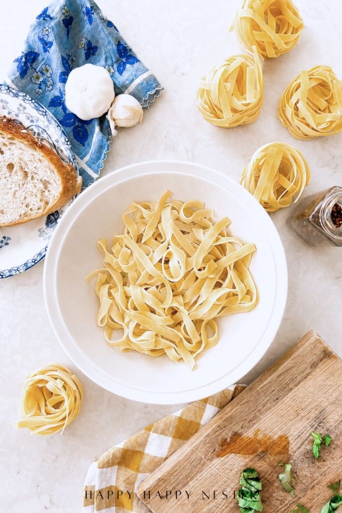 A white bowl of uncooked tagliatelle pasta sits on a countertop, surrounded by nests of pasta, a slice of bread, garlic, a blue-patterned napkin, a cutting board with herbs, and a jar of chili flakes.