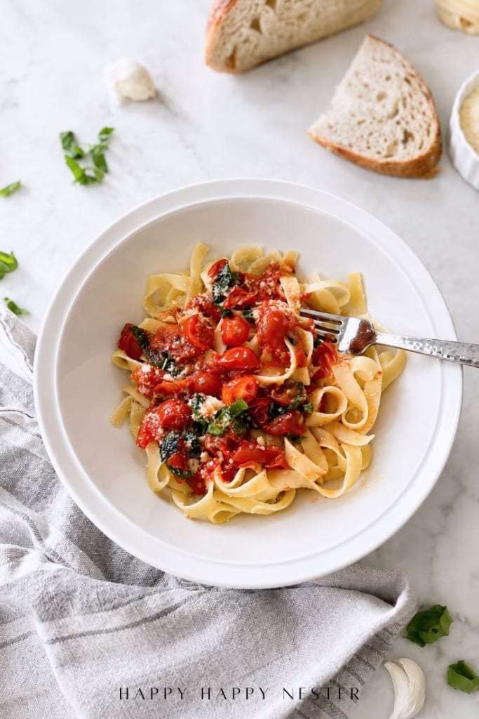 A white bowl of tagliatelle pasta topped with tomato sauce, cherry tomatoes, and fresh herbs, with a fork resting on the side. Slices of bread, garlic, and herbs are on a marble surface nearby.