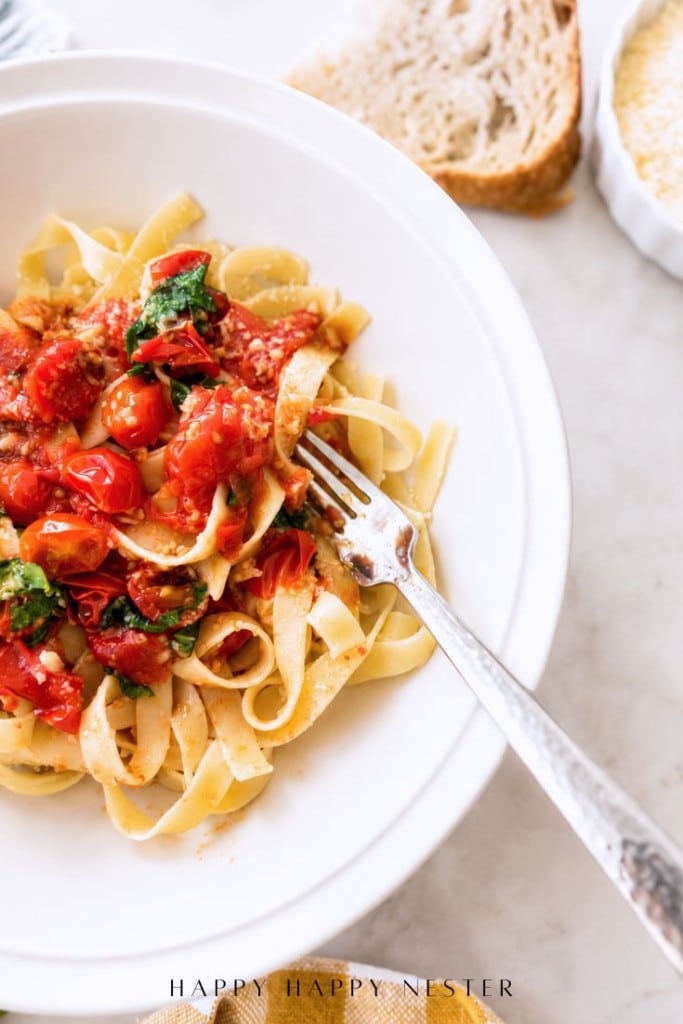 A white bowl of tagliatelle pasta topped with cherry tomatoes, fresh basil, and grated cheese, featuring a simple pasta sauce recipe; a fork twirls the pasta as slices of bread and parmesan sit in the background.