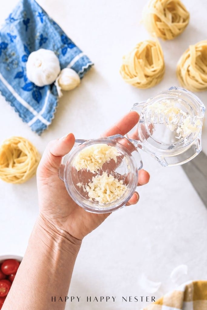 A hand holds a clear garlic press with freshly minced garlic inside, surrounded by pasta nests, a garlic bulb, a blue cloth, and cherry tomatoes on a light kitchen surface.
