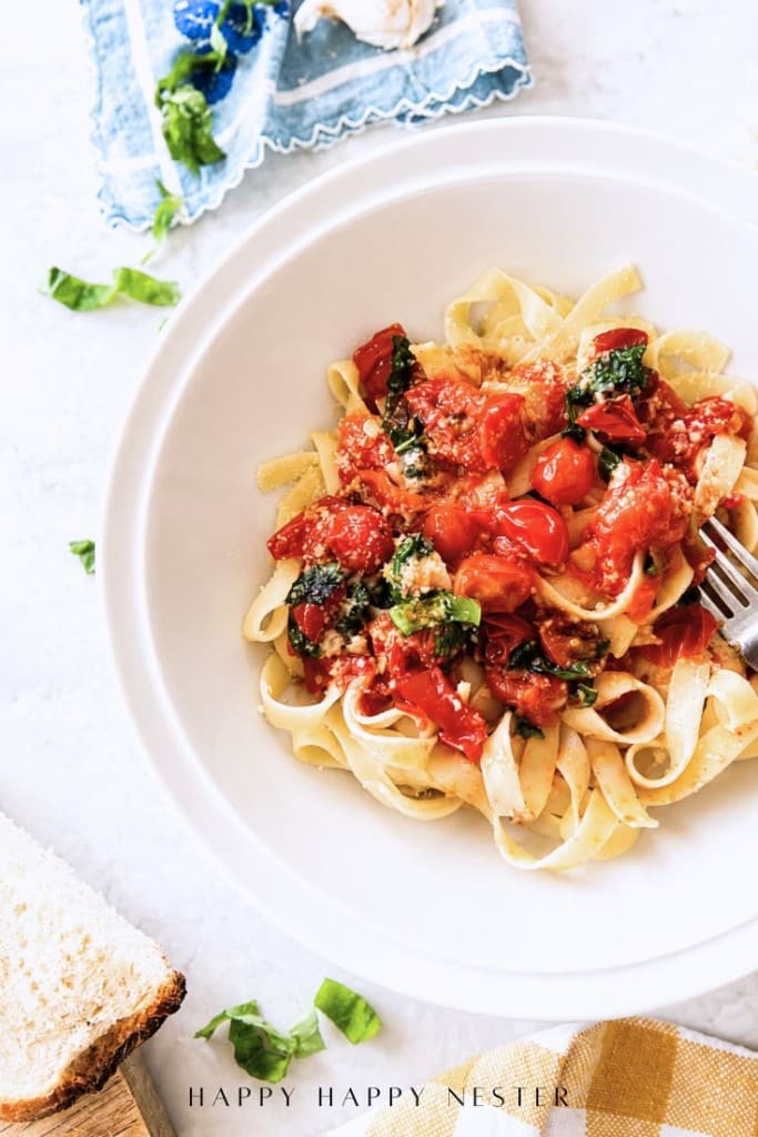 A white bowl of fettuccine pasta topped with a simple pasta sauce recipe of cherry tomatoes, basil, and grated cheese, with a fork on the side. Fresh basil, bread, and a napkin rest nearby on a light surface.