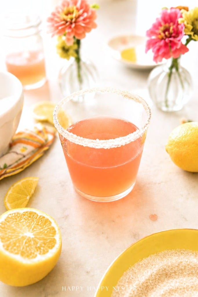 A glass of pink lemonade with a sugared rim sits on a light countertop, surrounded by lemon halves, a plate of sugar, and small vases with colorful flowers.