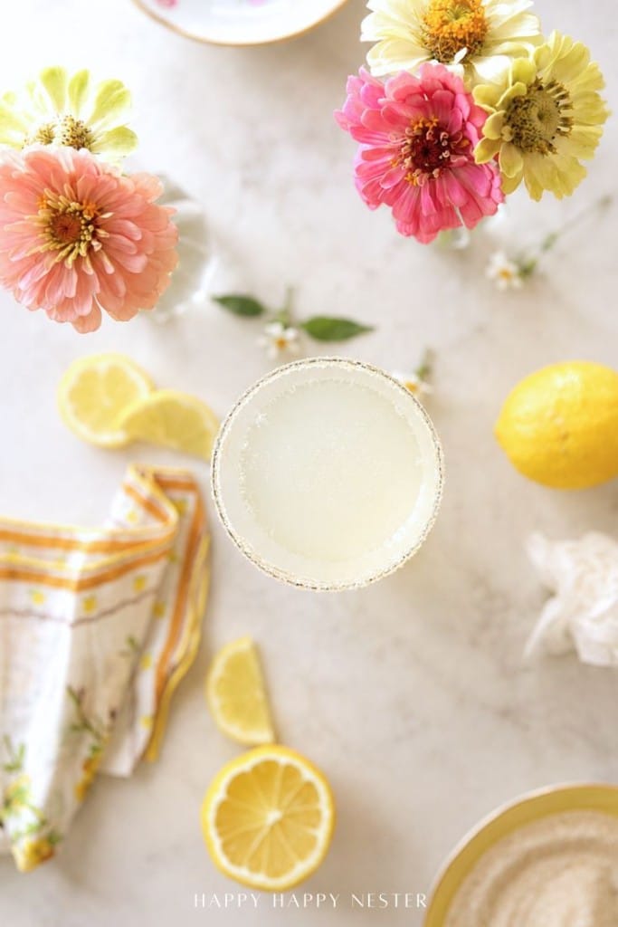 A cocktail in a glass with a sugared rim sits on a light surface, surrounded by lemon slices, a whole lemon, colorful flowers in vases, a yellow and white napkin, and a plate, creating a bright, cheerful scene.
