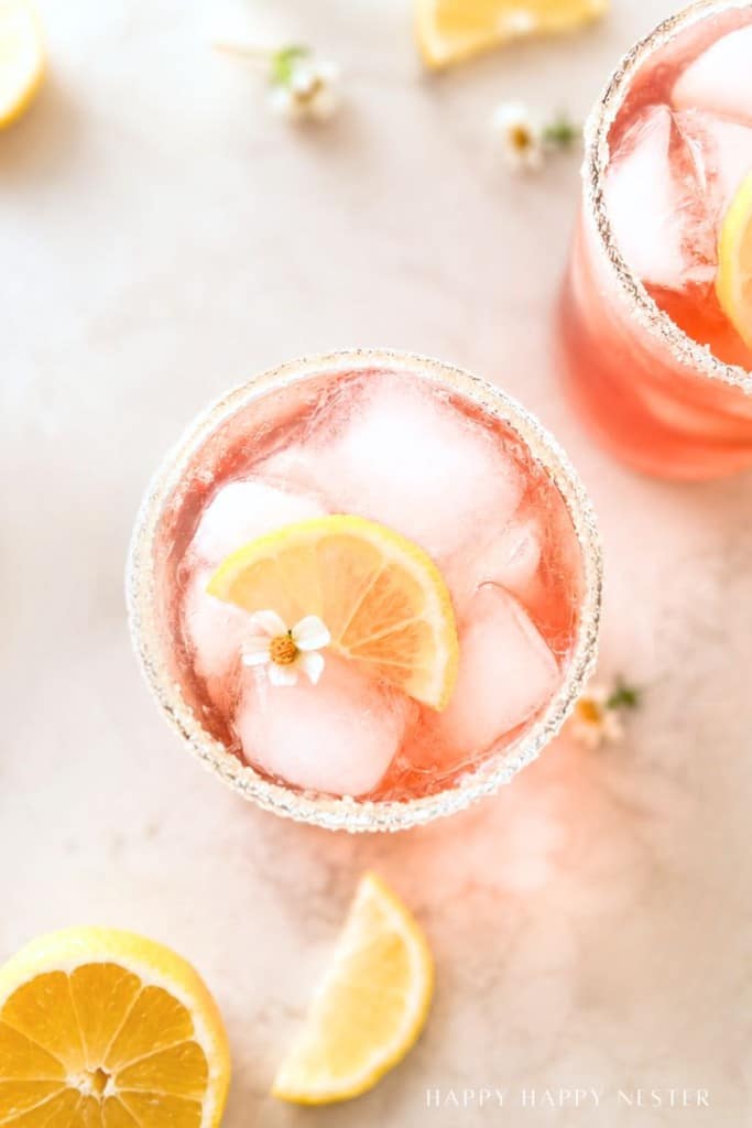 A glass of pink iced drink garnished with a lemon slice and small white flower, with additional lemon wedges and flowers scattered on a light surface nearby.