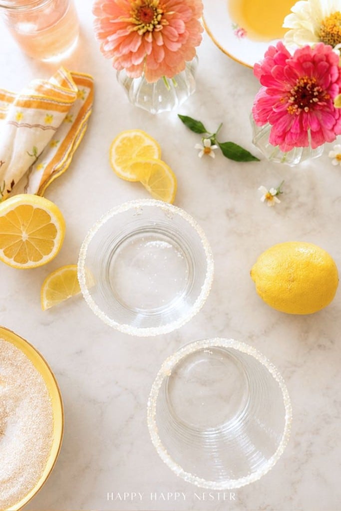 Two glasses with sugared rims sit on a marble surface surrounded by lemon slices, a whole lemon, a bowl of sugar, colorful flowers in jars, a yellow napkin, and a cup of tea.