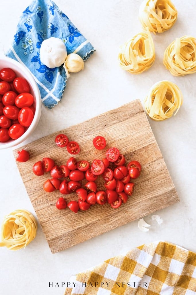 A wooden cutting board with sliced cherry tomatoes, surrounded by uncooked pasta nests, a bowl of whole cherry tomatoes, garlic, a blue cloth, and a yellow checkered towel on a white surface.