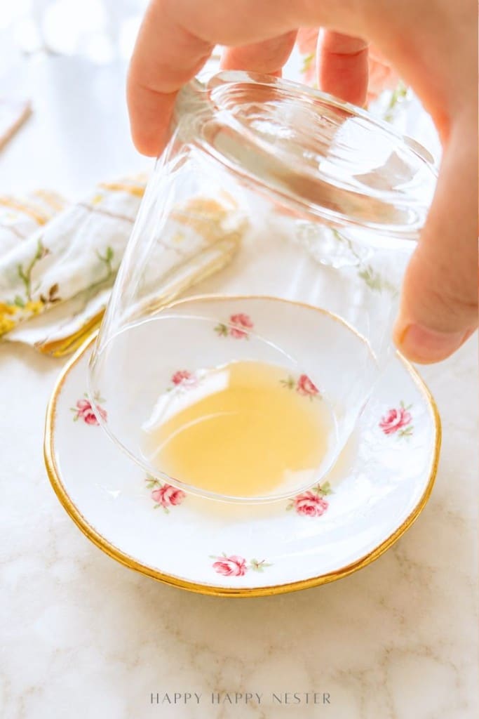 A hand holding an upside-down glass over a floral-patterned saucer with a small amount of light yellow liquid inside, on a marble surface with a napkin in the background.
