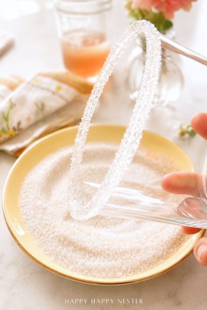 A hand rolls the rim of a clear glass in sugar on a yellow plate, with a floral napkin, drink, and flowers in the background—perfect prep for your favorite ginger beer mocktail recipe.
