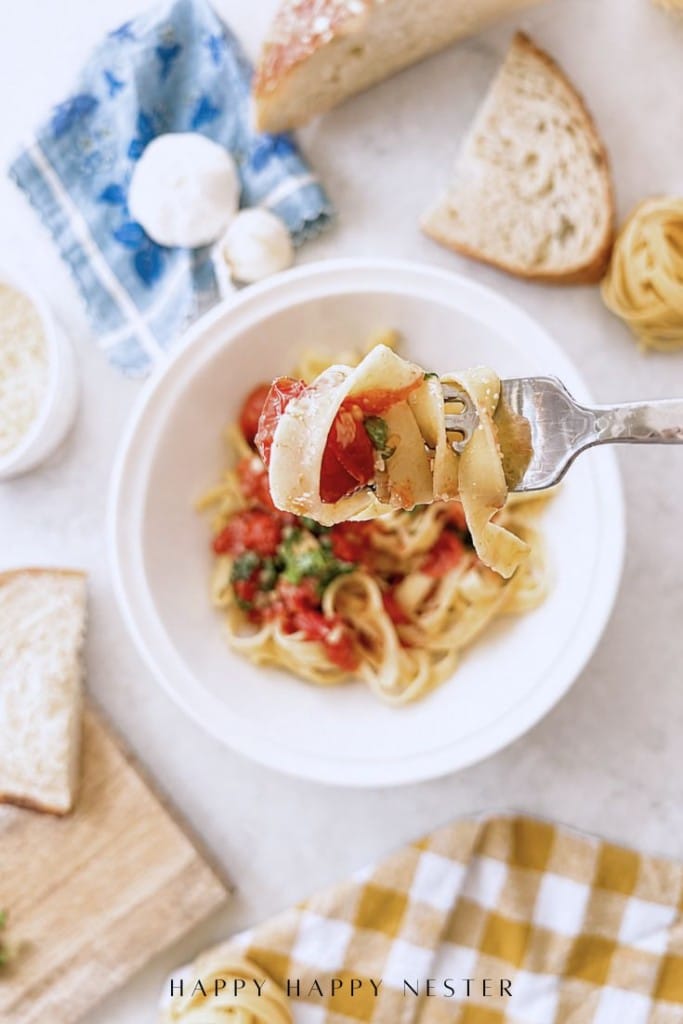 A fork holds a bite of pasta with tomatoes and basil above a bowl of pasta—a perfect showcase for your simple pasta sauce recipe. Slices of bread, a small cheese bowl, and a blue napkin rest on a light table nearby.