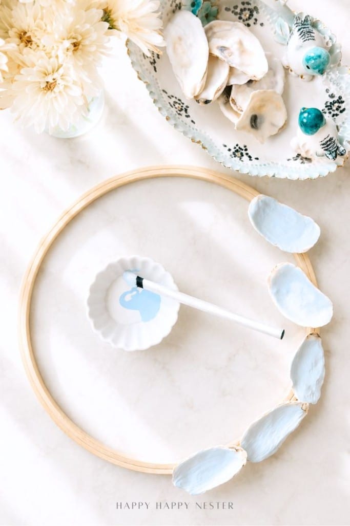 A wooden embroidery hoop with painted oyster shells attached is laid on a white surface, near a small dish with a paintbrush, a bowl of shells, turquoise beads, and cream flowers.