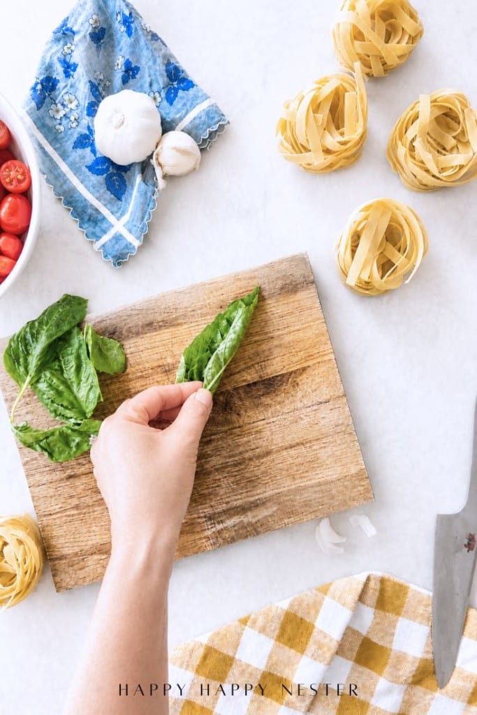 A hand rolls fresh basil leaves on a wooden cutting board surrounded by nests of uncooked pasta, a blue napkin with garlic and mozzarella, cherry tomatoes, and a yellow checkered towel.