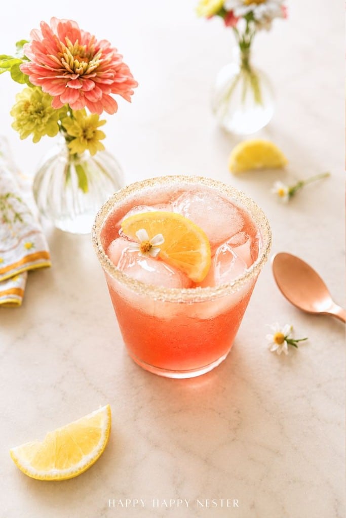 A pink iced ginger beer mocktail sits in a sugar-rimmed glass with a lemon slice and flower garnish on marble, surrounded by flowers, a lemon wedge, and a copper spoon.