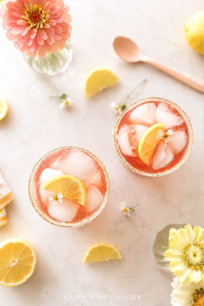 Two glasses of pink iced lemonade garnished with lemon slices and small white flowers, styled like a ginger beer mocktail recipe, are placed on a light surface with lemon wedges, flowers, and a gold spoon nearby.