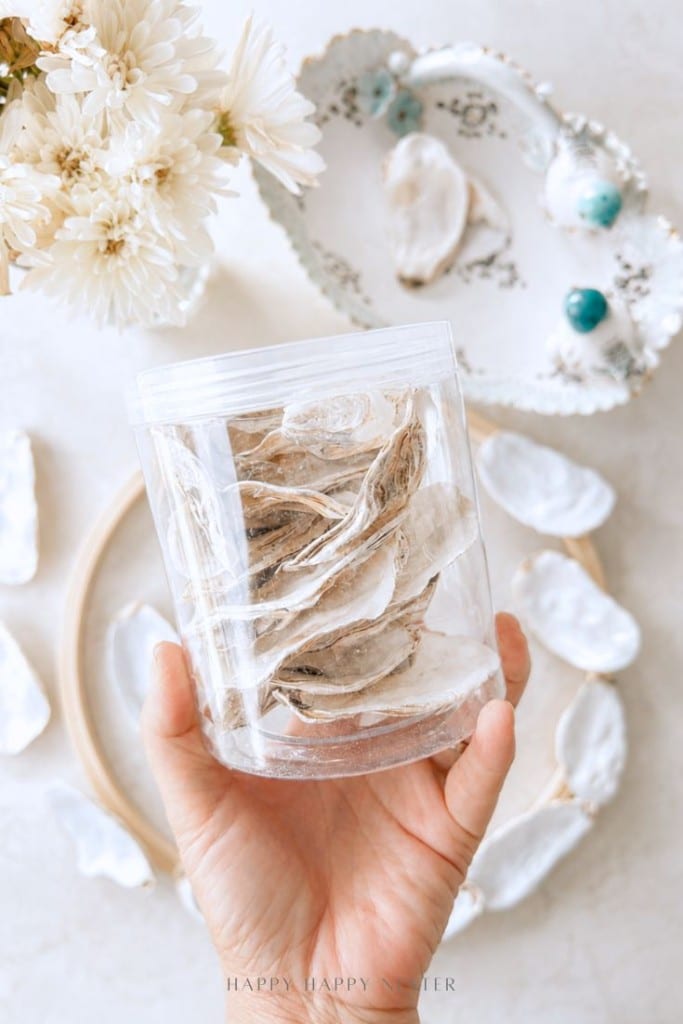 A hand holds a clear container filled with white oyster shells—perfect for your next oyster shell wreath DIY. More shells, a decorative plate, and a bouquet of white flowers rest on the light-colored surface in the background.