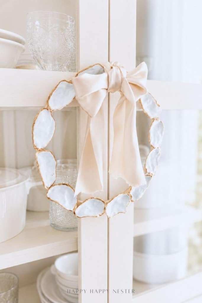 A decorative wreath inspired by an oyster shell wreath DIY, made of oval-shaped white stones with gold edges and topped with a large beige satin bow, hangs on a light-colored cabinet filled with glassware and dishes.