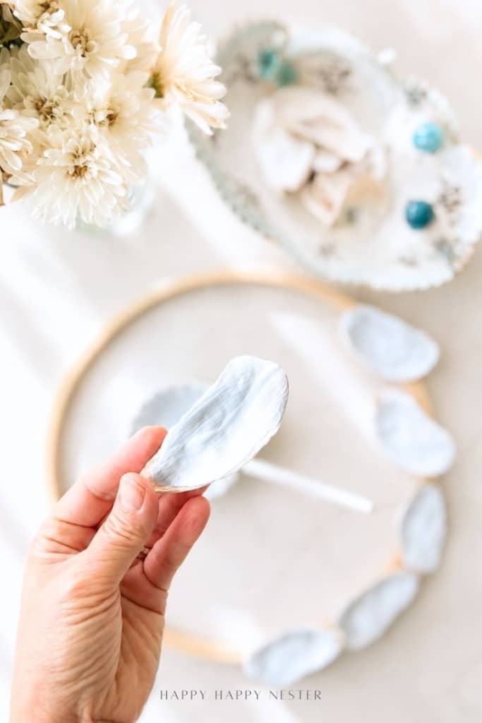 A hand holds a painted oyster shell above a table with white flowers, a bowl of shells and teal beads, and a wooden hoop decorated with similar painted shells.